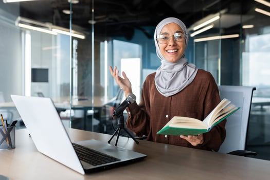 Online teaching. Young Muslim woman in hijab teacher sits at desk in office in front of laptop and microphone, holds book in hands. Conducts distance education. photo