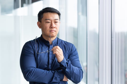 Portrait of a serious young Asian businessman, lawyer. He stands thoughtfully in the office by the window, holds glasses in his hands, looks to the side. photo