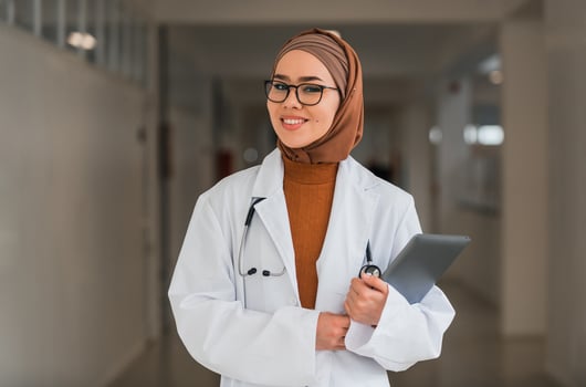 Portrait of happy Muslim female doctor or nurse wearing white coat with stethoscope in hospital corridor holding digital tablet looking at camera and smiling. Professional medical staff headshot. photo