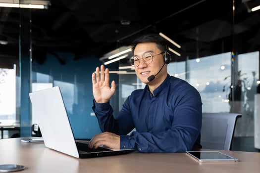 Successful smiling man working inside office using laptop and headset for call, asian cheerfully consulting tech support customer, businessman at work in shirt and glasses. photo