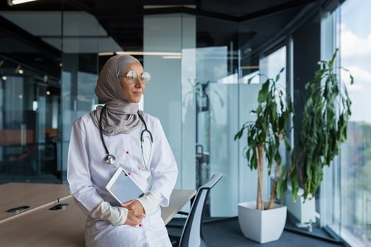 A happy and thoughtful young Muslim woman, an Arab doctor in a hospital, is sitting at her desk, holding a tablet in her hands, looking out the window, smiling, loving her work. photo