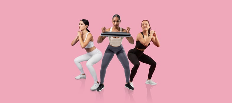 Three women engage in a squat exercise, showcasing their fitness routines in stylish workout attire. The bright pink background adds a vibrant touch to their focused activity. photo