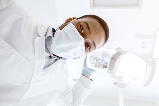 Low Angle View Of Black Stomatologist Turning On Lamp Before Check Up With Patient, Professional African American Dentist Doctor In Medical Mask Looking At Camera, Ready For Making Treatment photo