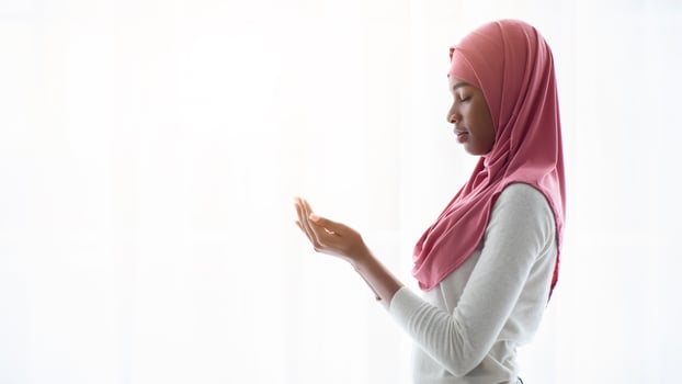 Portrait of young black muslim woman praying near window at home, religious lady in headscarf standing with closed eyes, making islamic pray dua gesture with cupped hands, side view, copy space photo