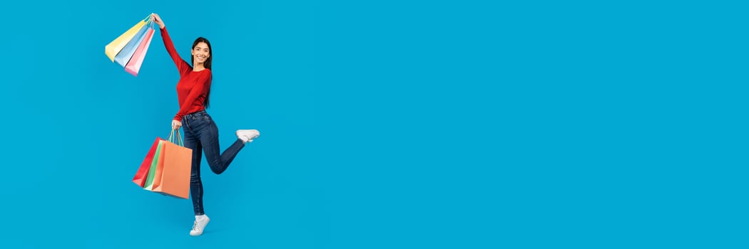 Excited young woman joyfully jumping in the air while holding shopping bags, showcasing her excitement for her purchases, posing on blue background, copy space photo
