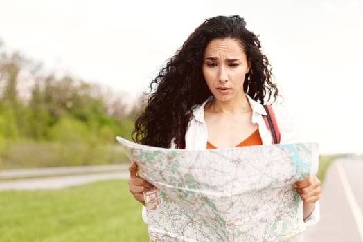 Confused millennial lady standing on road with map, feeling lost while choosing travel destination, hitchhiking for ride outdoors. Upset young woman traveling by autostop, cannot find correct route photo