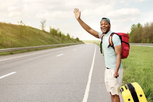 Happy black guy with backpack and suitcase flagging down car, needing ride, hitchhiking on roadside, copy space. Funky African American man traveling with luggage, having autostop journey, outdoors photo