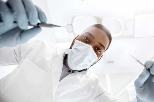 Black male dentist doctor with sterile dental tools in hands, low angle view. Portrait of african american stomatologist in protective medical mask checking up patient's teeth in clinic, closeup photo
