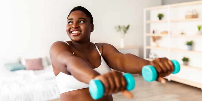 Smiling curvy young black woman doing exercises with dumbbells, during domestic weightloss training. Plus size millennial lady working out biceps muscles, staying healthy at home photo