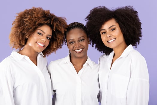 Three African American women stand close together, smiling warmly at the camera. They showcase their natural hairstyles and wear white tops, radiating friendship and joy in a modern setting. photo