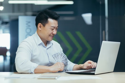 Happy young Asian businessman working in office on laptop and holding cash money banknotes in hands. photo