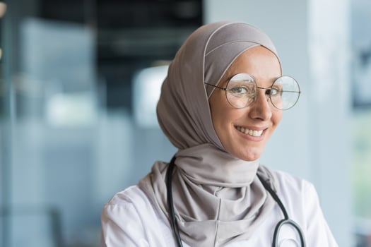 Close-up photo portrait of Muslim female doctor in gray hijab, female doctor smiling and looking at camera, female worker in glasses and white medical coat and glasses working inside modern clinic office