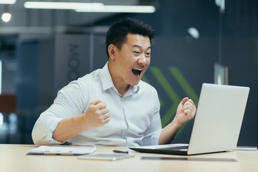 Overjoyed businessman celebrating success and victory, holding hands up, shouting with joy into laptop, asian man at work in modern office. photo
