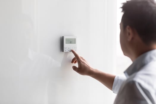 Close-up photo. The hand of a young man in a white shirt turns on the control buttons of the air conditioner hanging on the wall. Standing on the right photo