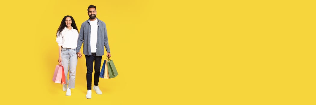 Big Sales. Full Length Of Happy Arab Couple Walking With Shopping Bags In Studio On Yellow Background. Smiling Young Middle Eastern Man And Woman Holding Hands Carrying Purchases, Copy Space photo