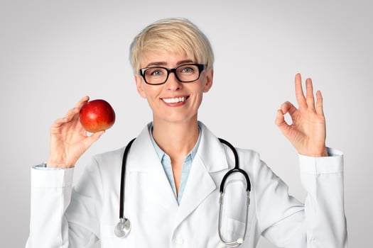 Diet, healthy eating and doctors advice. Smiling female doctor nutritionist in glasses and white coat holds red apple and shows okay sign with hand isolated on blue background, free space, studio shot photo