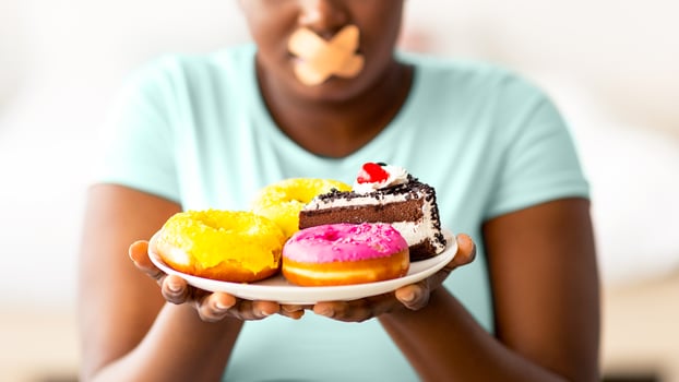 Diet restrictions. Curvy black woman with adhesive bandage on her mouth looking at plate of sweets in hunger at home. Plus size African American lady refusing to eat junk food, losing weight photo