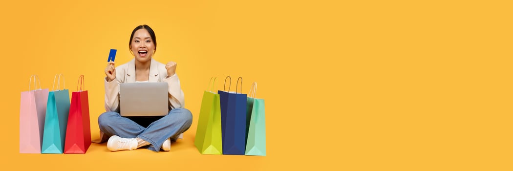 Glad excited millennial chinese lady with shopping bags, credit card, laptop, enjoy sale and win, isolated on orange studio background. Finance for purchases, shopaholic emotions, cashback, copy space photo