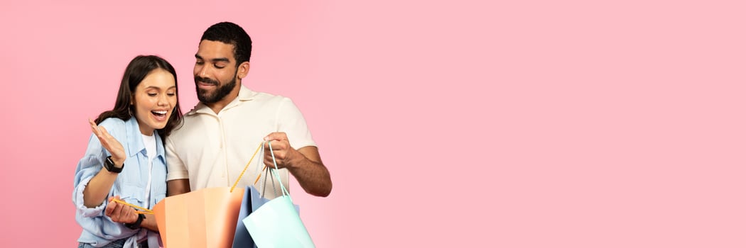 Excited happy pretty young woman looking inside colorful paper bags, shopping with her boyfriend or husband during black friday, season sale, pink background, copy space. Retail, consumerism photo