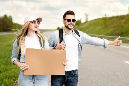 Happy millennial couple standing along highway with empty sign, stopping car, having road trip, mockup for design. Wanderlust, autostop journey, hitchhiking together, summer freedom concept photo