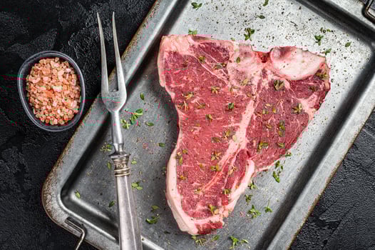 T-bone steak, raw marbled beef meat in a steel kitchen tray with herbs. Black background. Top view photo