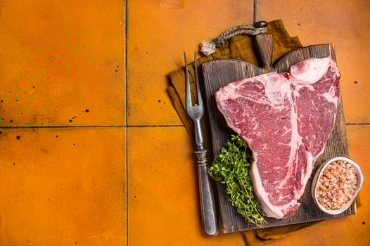 Porterhouse raw beef meat Steak, T-bone with thyme and sea salt on a wooden board. Orange background. Top view. Copy space photo