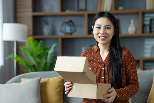 Asian woman with long black hair opening a cardboard package, smiling. She enjoys a moment of happiness in a home setting, surrounded by cozy decor, capturing joy of unboxing. photo