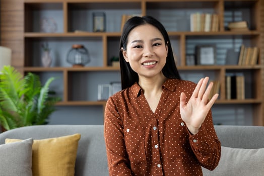 Asian woman sitting on sofa waving warmly at camera. She's wearing a brown polka dot shirt, expressing friendliness and happiness. Ideal for virtual meetings, online communication, home setting. photo