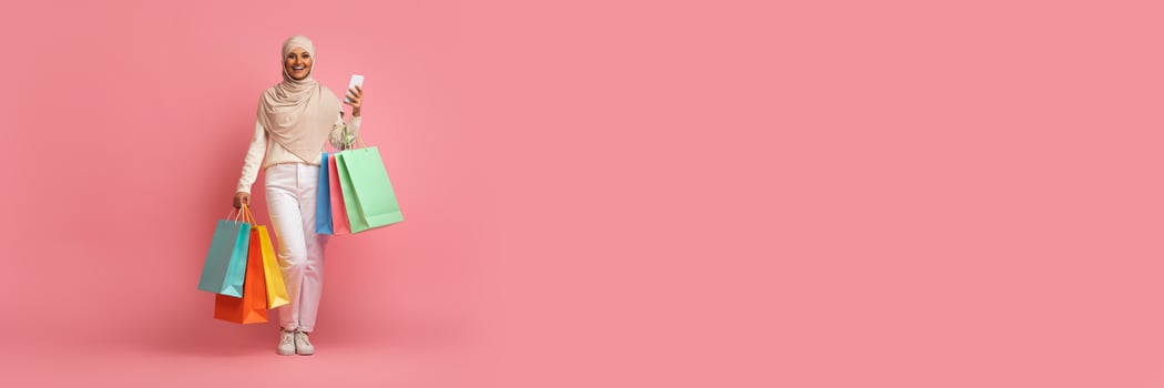 Muslim woman dressed in casual attire holds multiple shopping bags while smiling happily. She enjoys her shopping experience during a sale, with a bright pink background enhancing the festive mood. photo