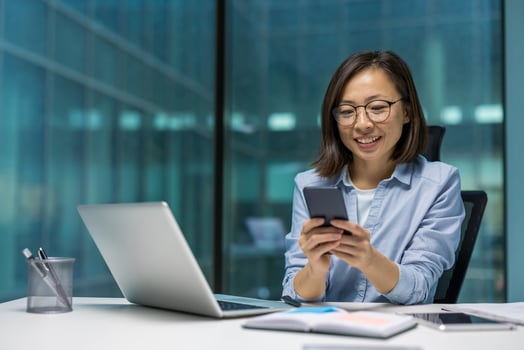 Asian businesswoman using smartphone at desk, working on laptop, smiling in modern office. Highlights digital productivity, communication, technology. Ideal for business, work themes. photo