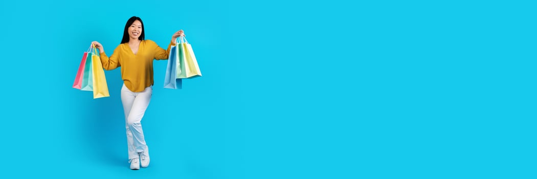 Successful shopping. Positive cheerful young chinese lady wearing casual outfit showing colorful shopping bags and smiling at camera, blue studio background, copy space for ad photo