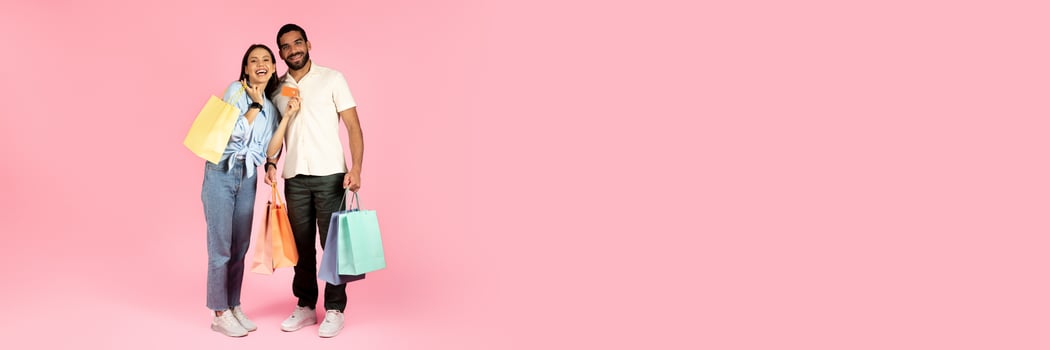 Happy cheerful beautiful young couple shopping together on pink studio background, handsome guy and pretty lady holding colorful paper bags, purchases and bank card, copy space photo