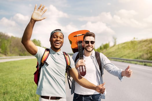Two male friends with backpacks and guitar hitchhiking on highway, trying to catch free ride, traveling together on summer vacation. Multiracial guys having autostop journey, standing on roadside photo