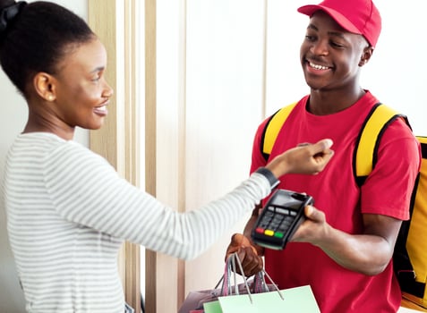 Cheerful Black Female Paying Courier Putting Smartwatch To Terminal Receiving Shopper Bags Standing At Home. Modern Shopping And Delivery Service. Selective Focus photo