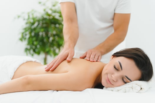 Woman enjoying professional body massage at a modern spa salon. Relaxed young lady lying on a massage table while a skilled masseur is gently massaging her back. Beauty, health, pleasure, concept photo