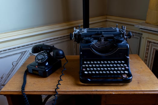 A black typewriter sits on a wooden desk next to a black rotary phone photo