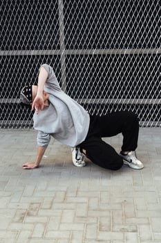 A young man wearing a gray sweatshirt and black pants is performing a dance move in front of a wire fence photo