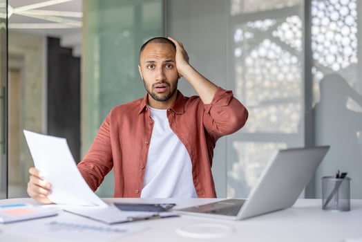 A professional looks worried while examining documents in a modern office setup, showing stress or confusion. The setting includes a laptop and charts, highlighting a business or financial context. photo
