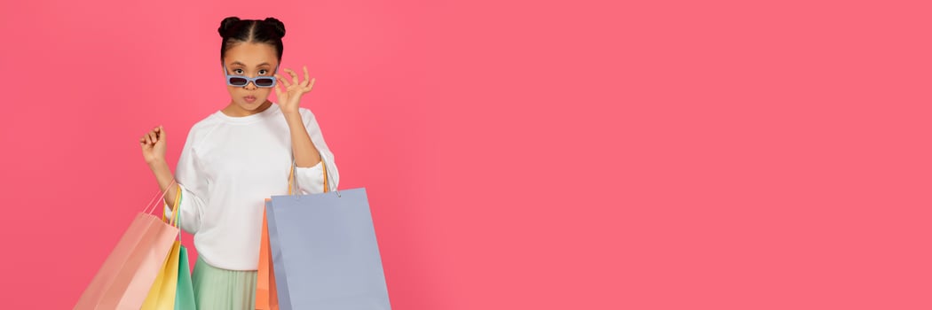Stylish asian woman with shopping bags adjusts sunglasses and looking at camera, young korean shopaholic lady carrying purchases, enjoying fashion and sales, standing on pink background, copy space photo