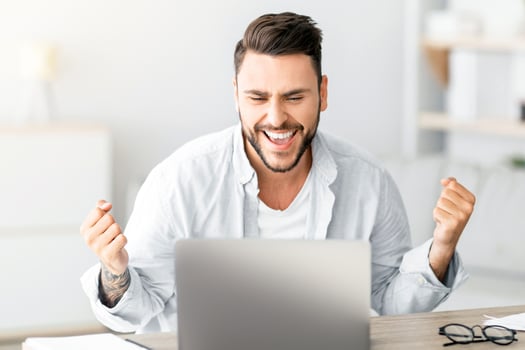 Portrait of overjoyed guy celebrating success, shaking clenched fists while sitting at desk in living room interior, copy space. Man using laptop computer, reading great news, screaming yes photo