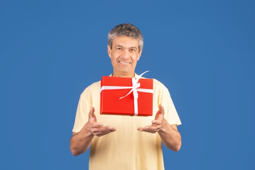 A middle aged man in his 50s is cheerfully presenting a red gift box tied with a white ribbon. He stands alone against a solid blue backdrop, showcasing excitement and joy. photo