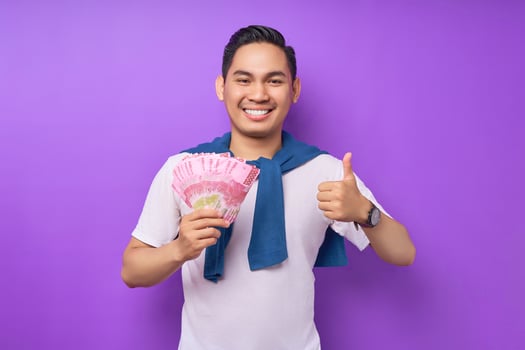 Smiling young Asian man wearing white t-shirt holding cash money in rupiah banknotes and gesturing thumbs up isolated over purple background. people lifestyle concept photo
