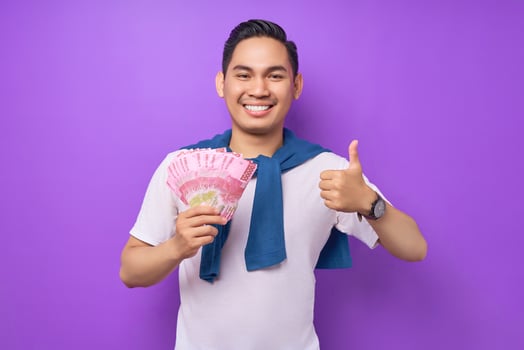 Smiling young Asian man wearing white t-shirt holding cash money in rupiah banknotes and gesturing thumbs up isolated over purple background. people lifestyle concept photo