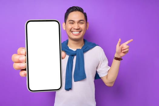Cheerful young Asian man holding smartphone and pointing aside at copy space isolated over purple background photo