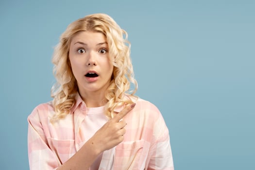 Shocked, amazed blonde teenager, with open mouth looking at camera, pointing finger on copy space photo