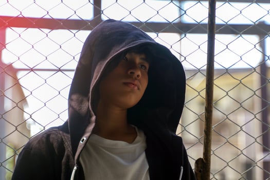 Asian teenage boy wearing a black hood over his head, standing with his back against a mesh fence, eyes looking down. Concept of backlit image, teenager, hacker. photo