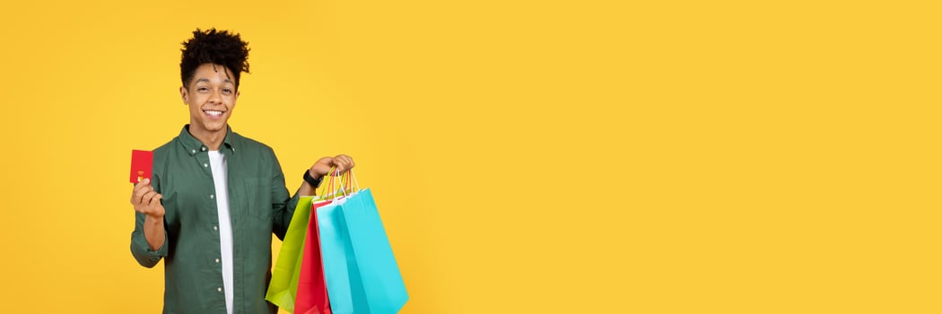 Positive stylish african american guy showing red plastic bank credit card and colorful paper bags purchases, isolated on yellow studio background. Easy shopping concept, copy space photo