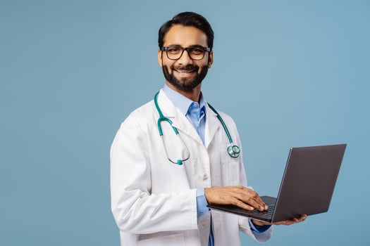 Smiling Indian man, doctor wearing stylish eyeglasses, white coat and stethoscope, holding laptop photo