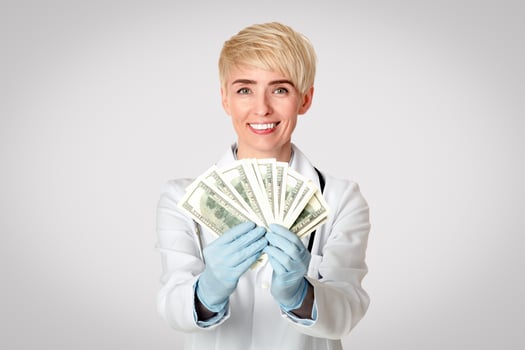 Corruption of medical system and taking bribe. Happy female doctor in white coat and rubber gloves received money and shows dollars to camera, isolated on gray background, free space, studio shot photo