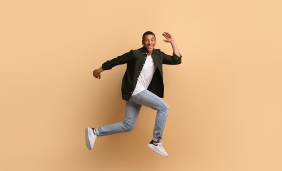 A joyful African American man is leaping in a dynamic pose. He wears casual attire with jeans and a shirt, showcasing an upbeat lifestyle while posed against a soft, beige backdrop. photo
